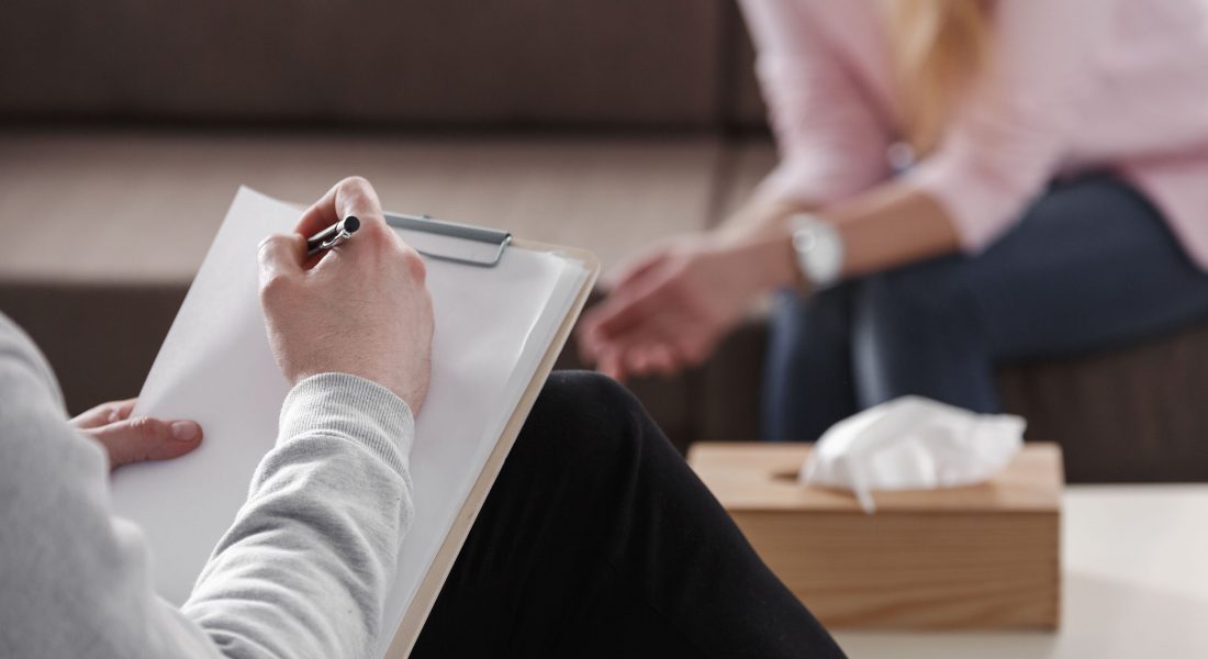Close-up of therapist hand writing notes during a counseling session with a single woman sitting on a couch in the blurred background.