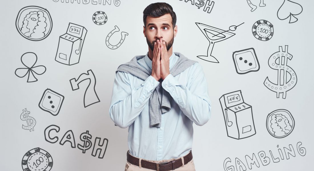Oops! Confused young bearded man is covering mouth with hands and looking away while standing against grey background with different doodle illustrations on it. Gambling concept. Face expressions